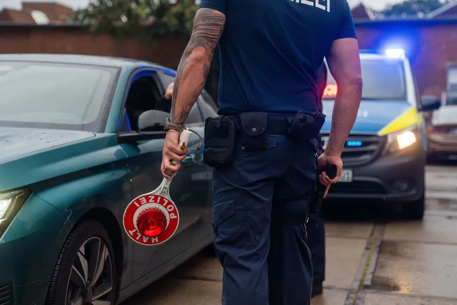 Polizist hält ein Stoppschild neben einem grünen Auto, im Hintergrund blinken Blaulichter